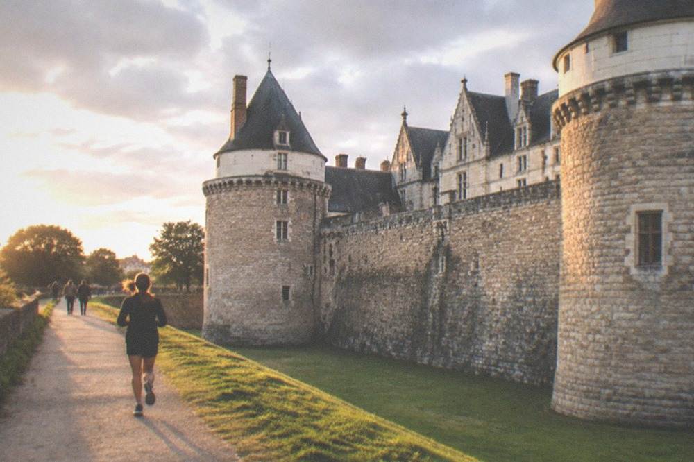 The dry moat and grassy rampart walk of the château with the towers of the fortress rising above and pedestrians on the path