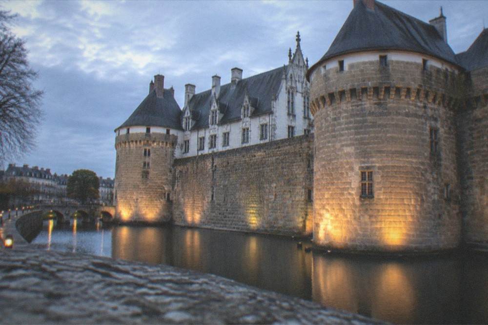 The floodlit ramparts and towers of the château seen at dusk with the walkway lamps glowing along the moat