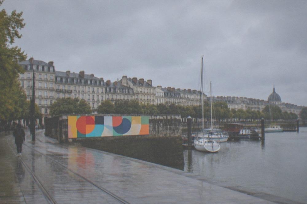A bright abstract artwork mounted on a harbour wall with moored boats behind it