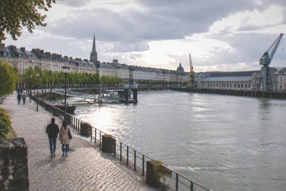 A couple walking along a riverside promenade towards large port buildings