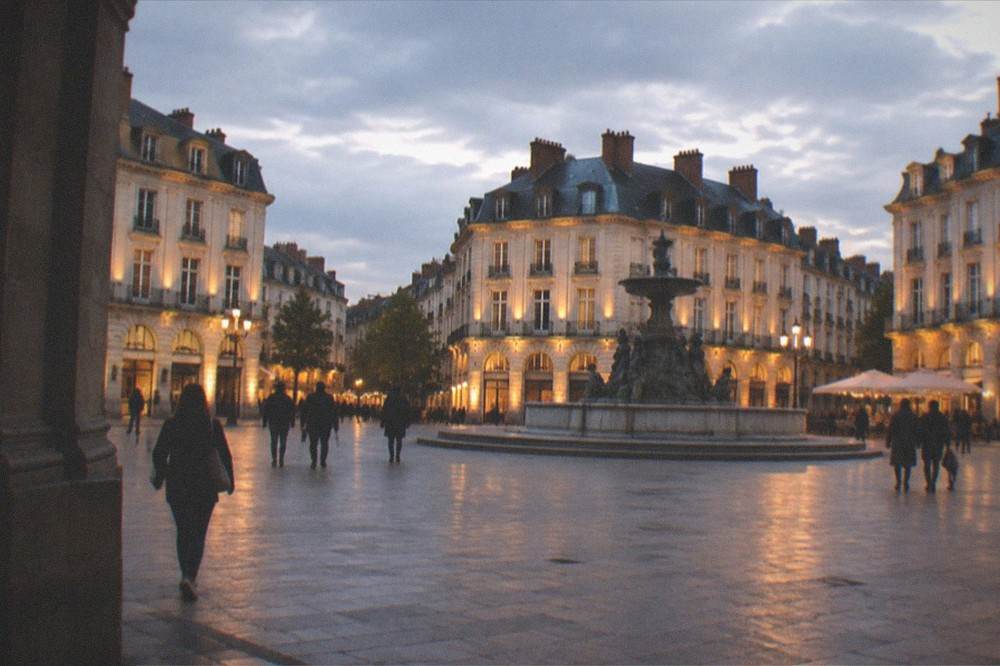 A wide city square at dusk with illuminated buildings and people walking