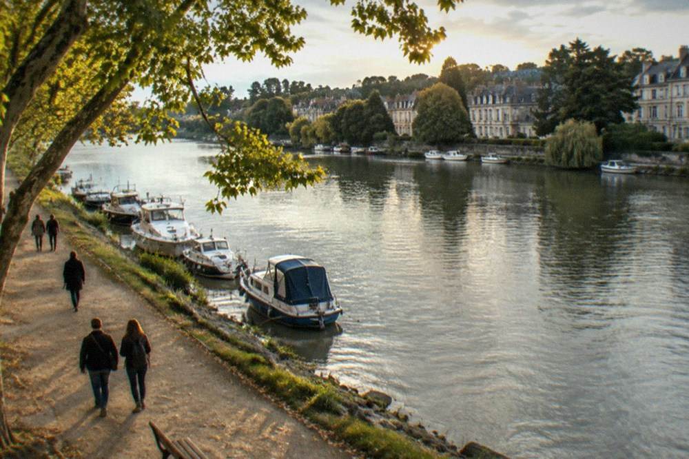 Visitors walking beneath plane trees along the quayside of the Erdre river in Nantes with moored boats on the water