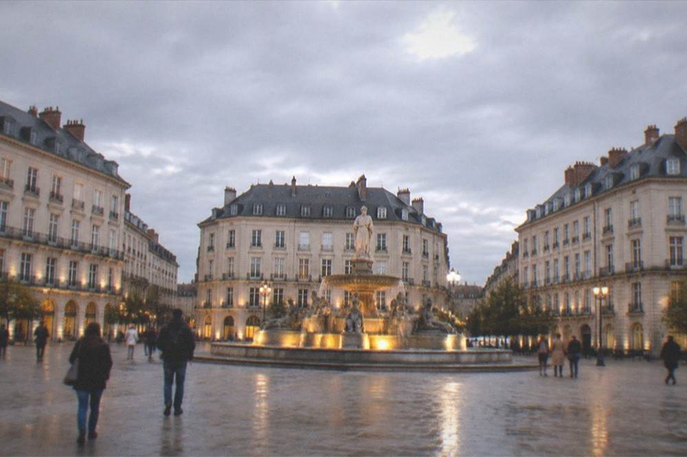 Evening view of a lit fountain and classical facades on Place Royale in central Nantes with people walking across the square