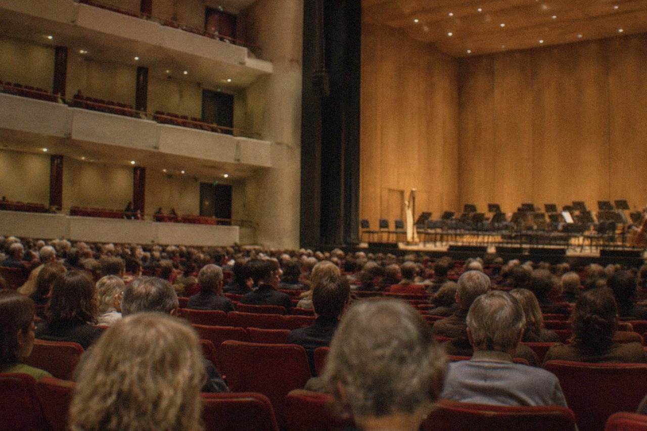 A concert hall audience seated in rows of red seats waiting for an orchestra to begin