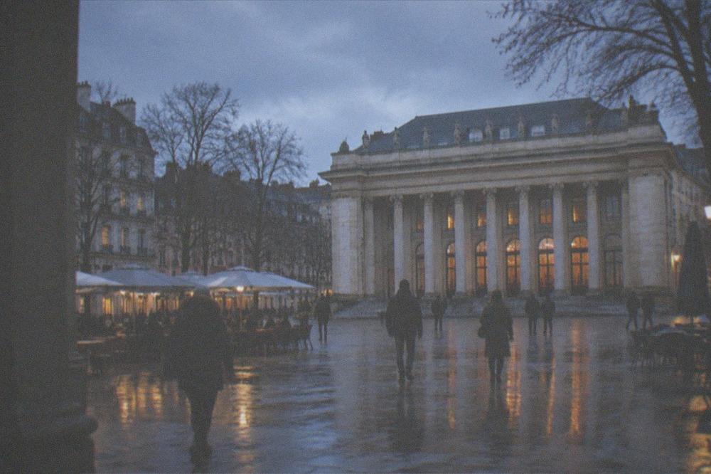 People in winter coats walking across a European city square under bare trees