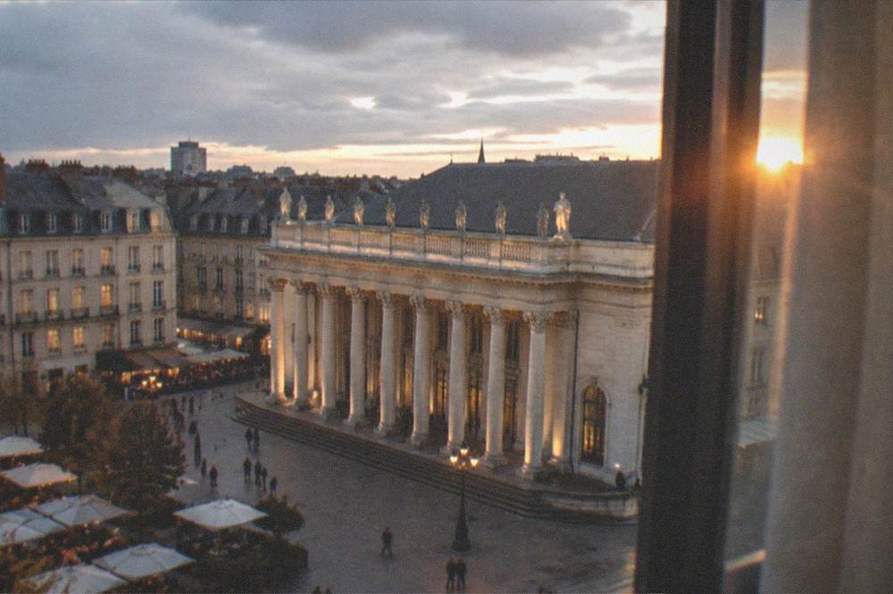 A hotel room window overlooking European rooftops at dusk with lights coming on