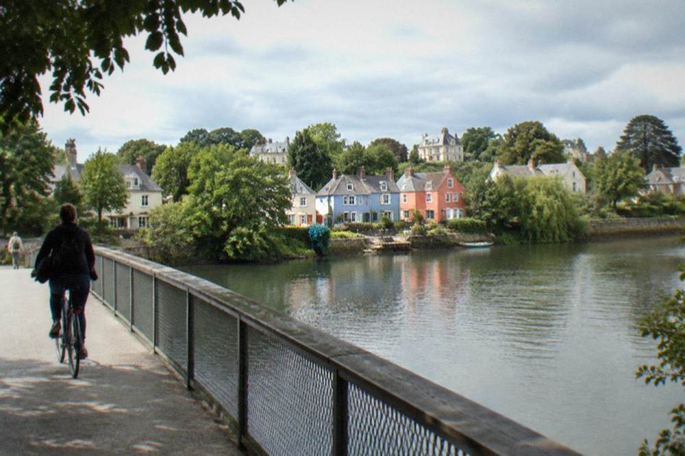 A cyclist crossing a bridge over a calm river toward colourful fishermen's houses on the far bank