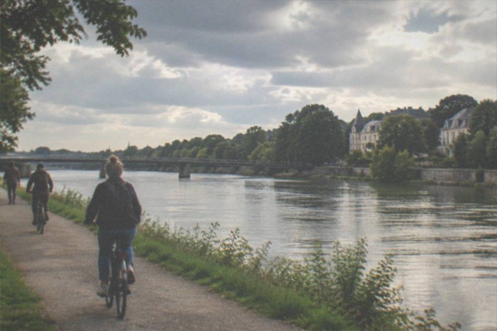 A wide river in late afternoon light with a long bridge and cyclists on a path near the bank