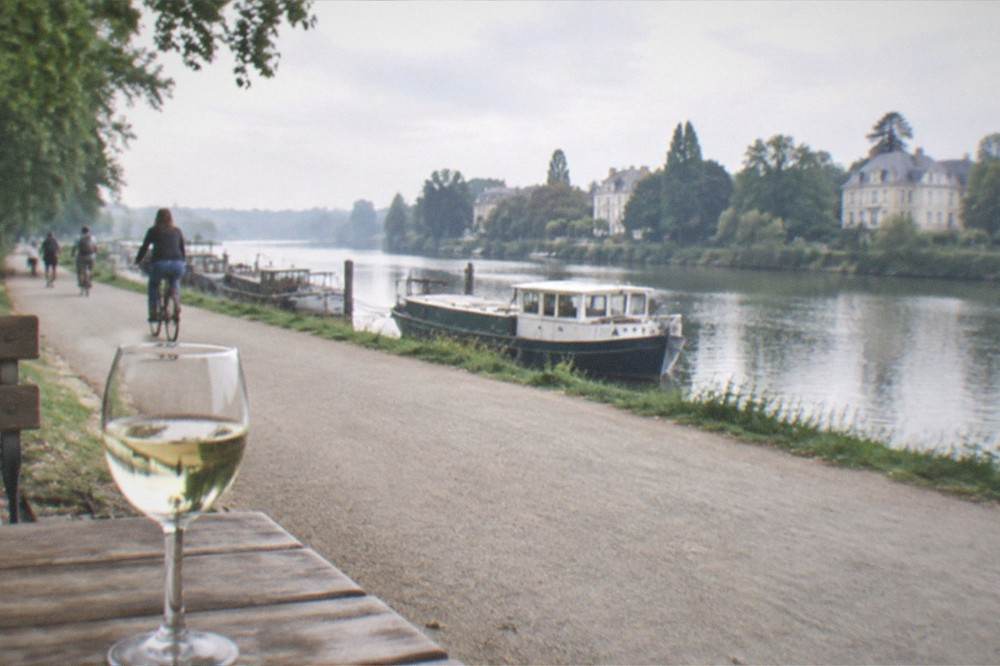 A glass of white wine on a wooden table beside a river with moored boats in the background