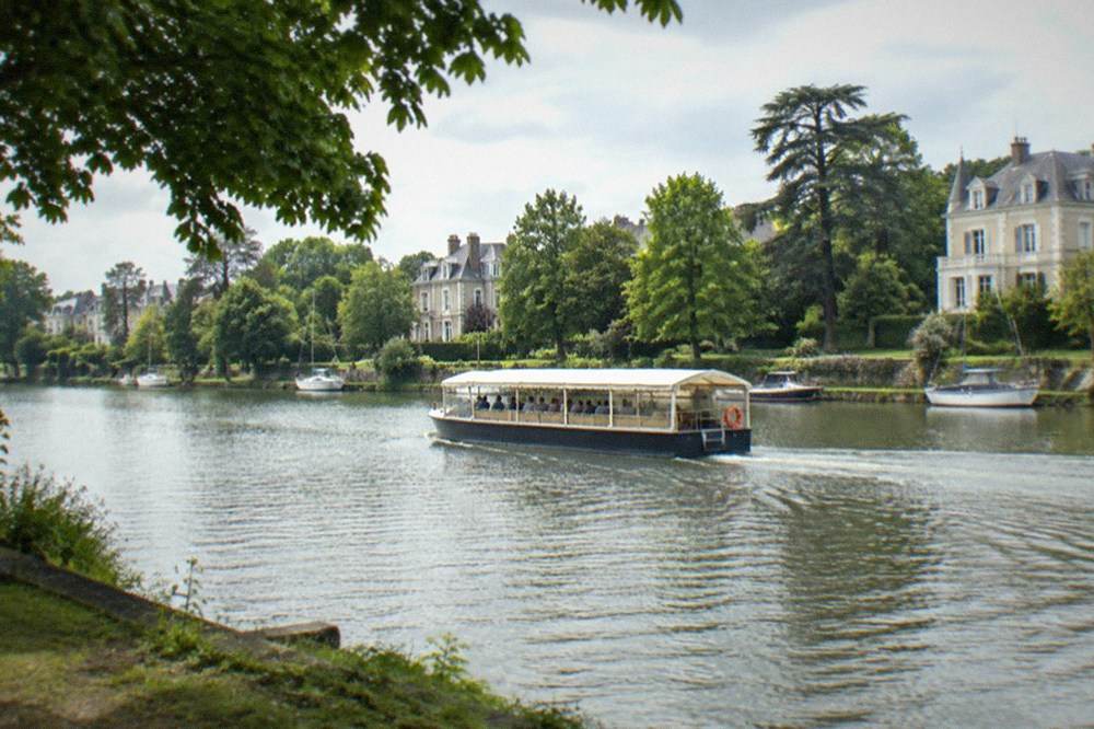 A covered sightseeing boat travelling along a narrow tree-lined river with lawns and grand houses on the far bank