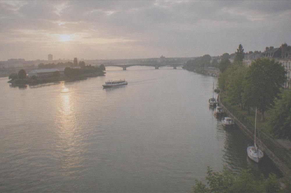 A wide river at golden hour with a bridge, an island with warehouses on the left bank and a ferry mid-crossing