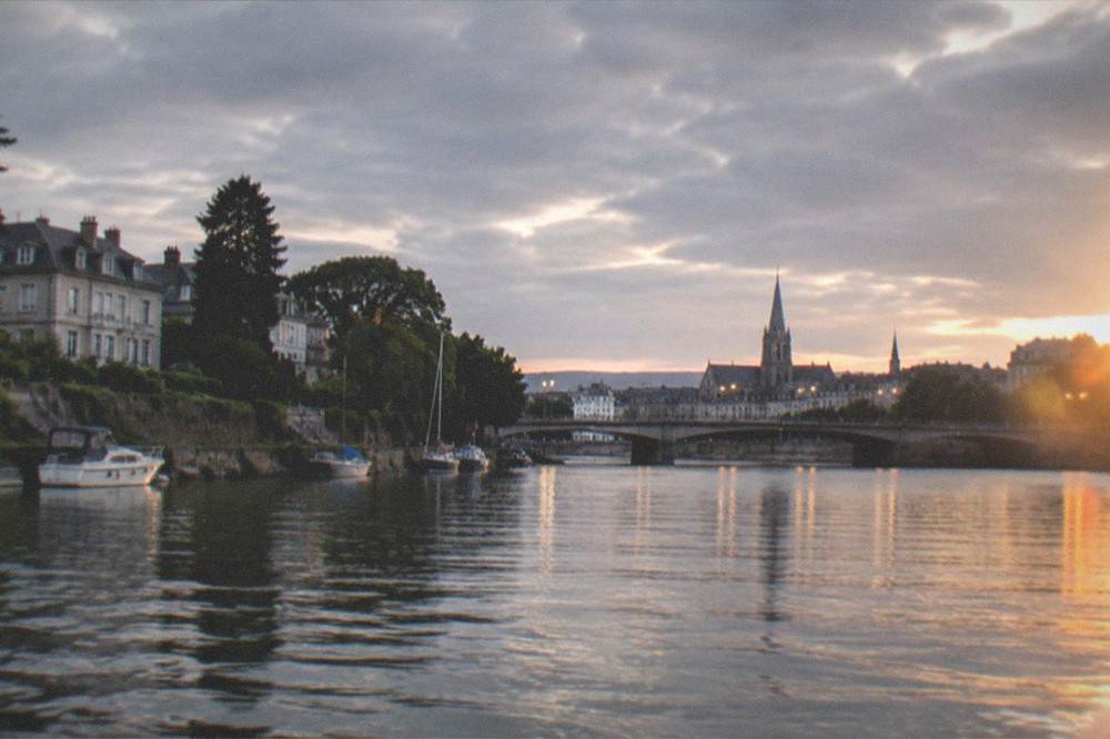 A riverfront at dusk seen from the water, with lit bridges crossing to a city centre of church spires and apartment buildings