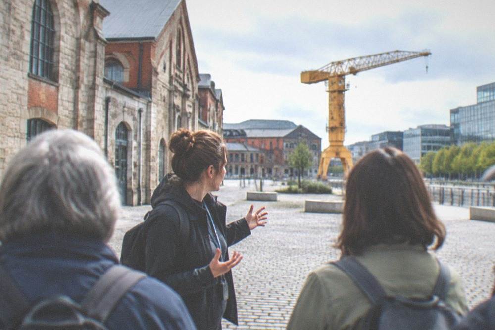 A guide speaking with a small group of visitors on a cobbled square beside old stone buildings