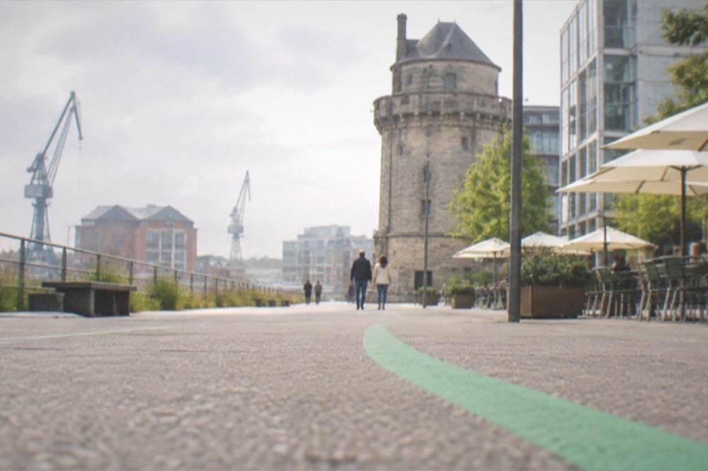A green painted line on a city pavement leading past a historic stone tower and modern café terraces
