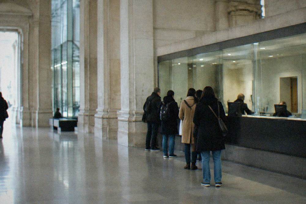 A museum ticket desk with a glass counter and visitors queueing in a stone entrance hall
