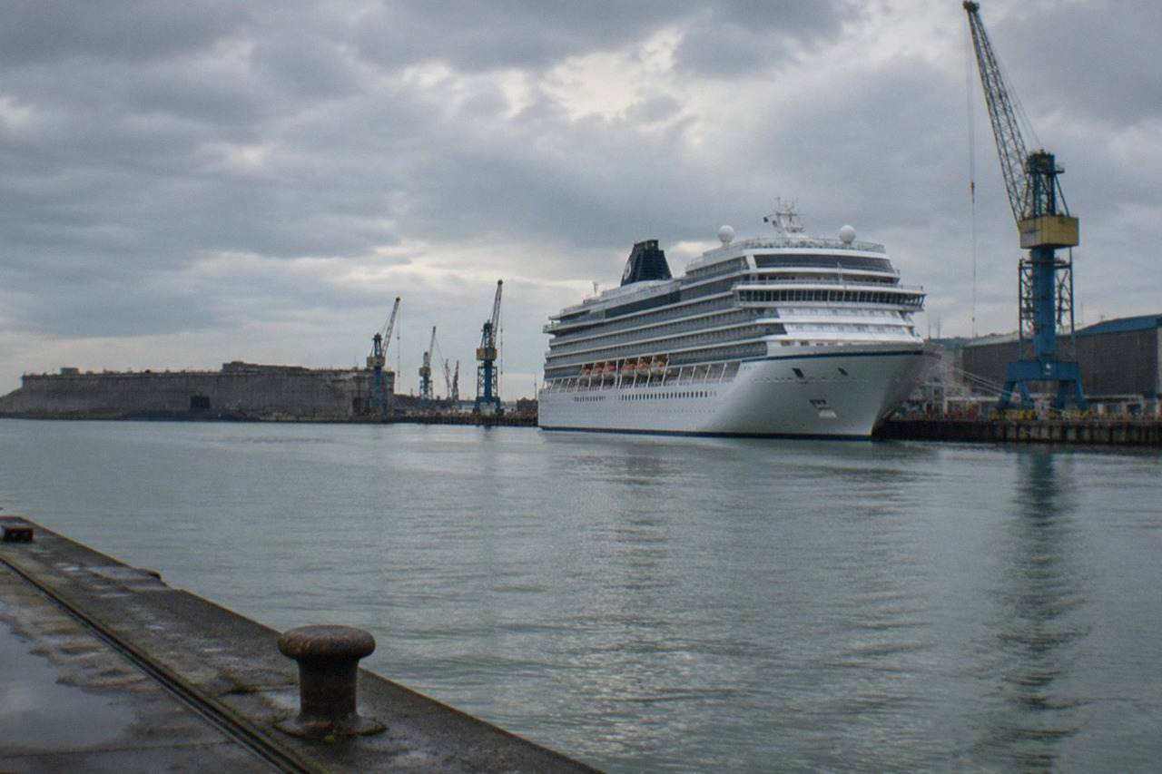A large cruise ship docked at a French Atlantic port terminal with cranes and warehouses in the background