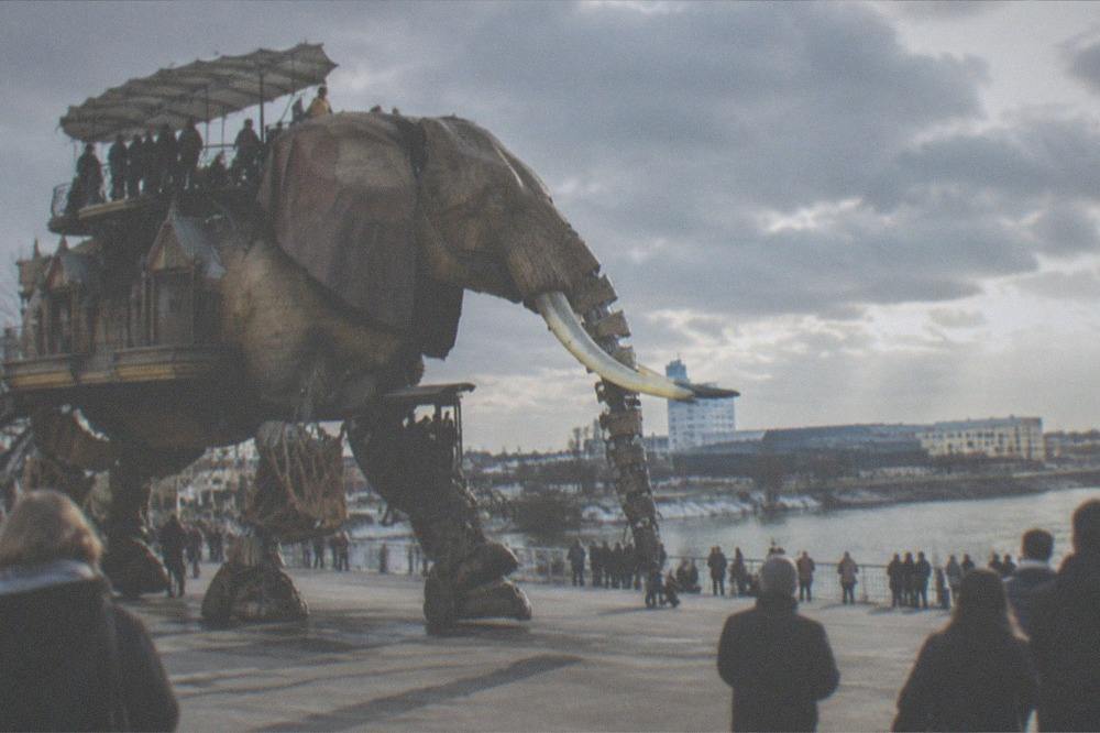 A giant mechanical elephant with wooden lattice legs carrying passengers along a waterfront promenade