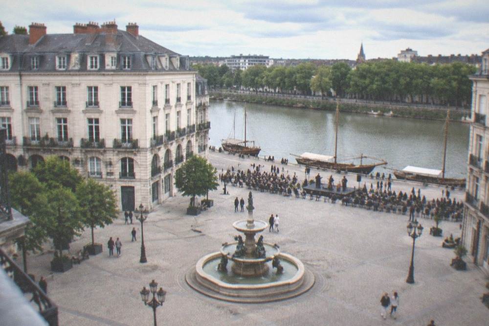 Classic wooden boats moored along a tree-lined river bank while people listen to a concert on the quay