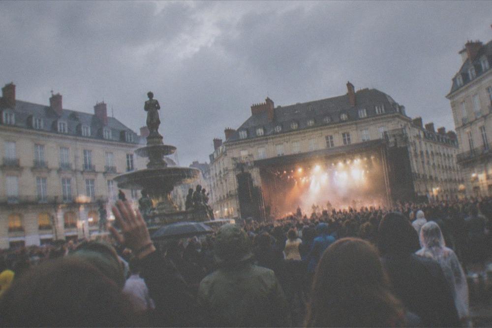 A festival crowd with raised hands in front of a large illuminated stage at dusk