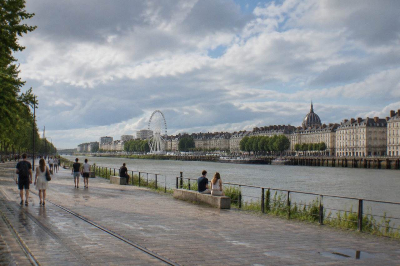 People strolling along a sunlit Loire riverside quay in Nantes with the ferris wheel and island skyline behind them
