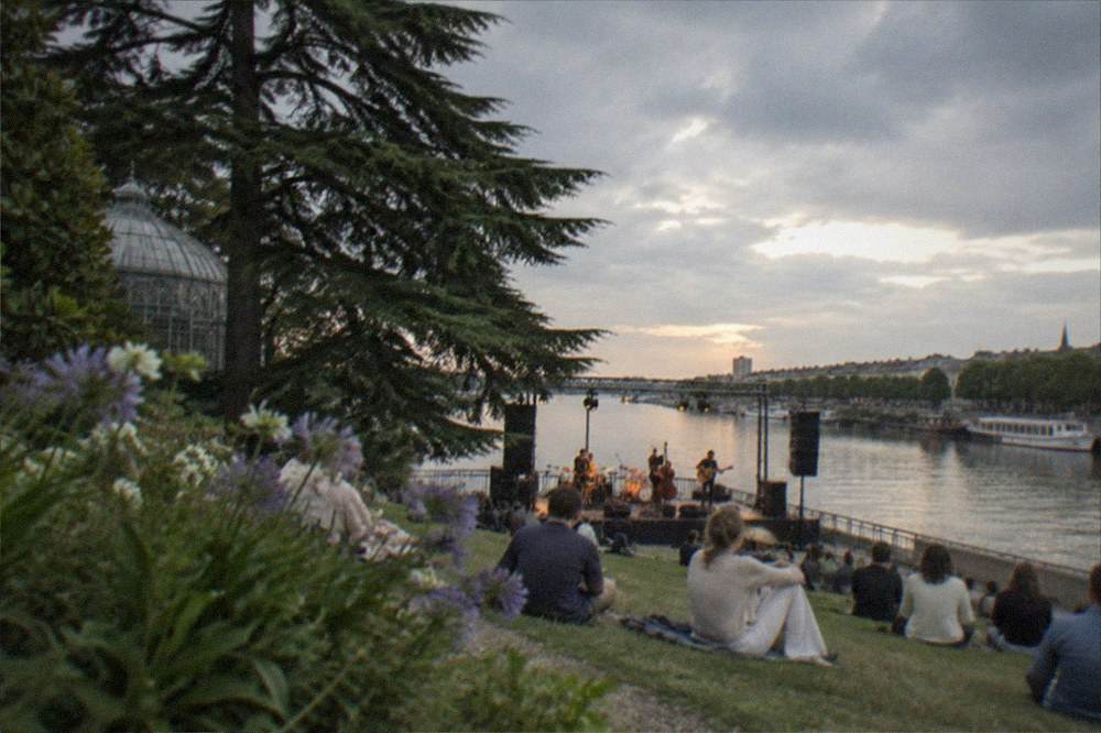 Musicians performing on an outdoor riverside stage at dusk with boats moored along the bank