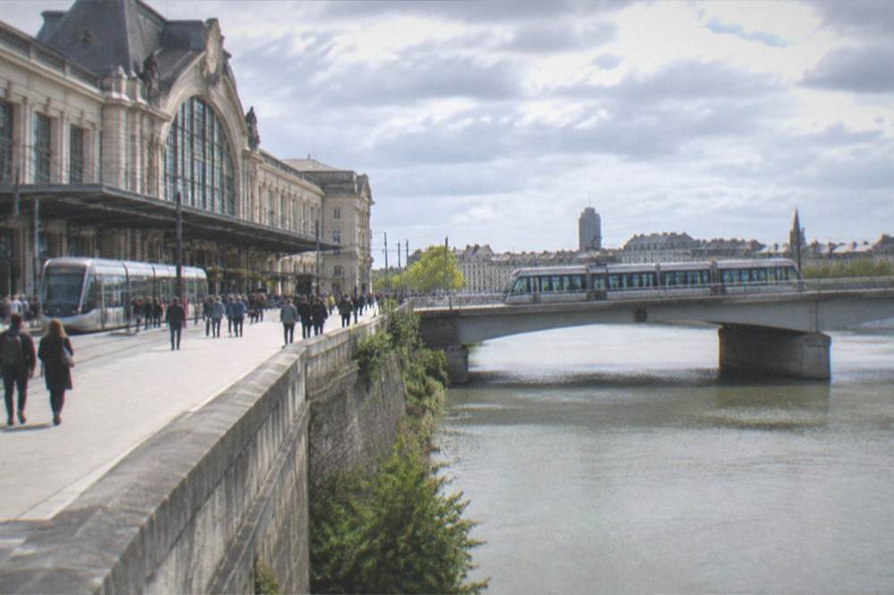 A tram crossing a bridge over a wide river with a city skyline beyond