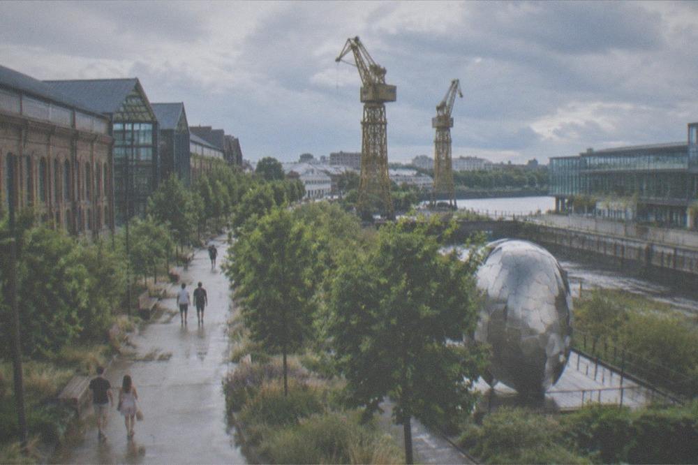 Visitors walking a tree-lined city path in warm summer light beside a public artwork