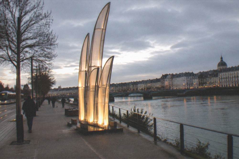 An evening view of a lit public artwork beside a riverfront promenade in Nantes