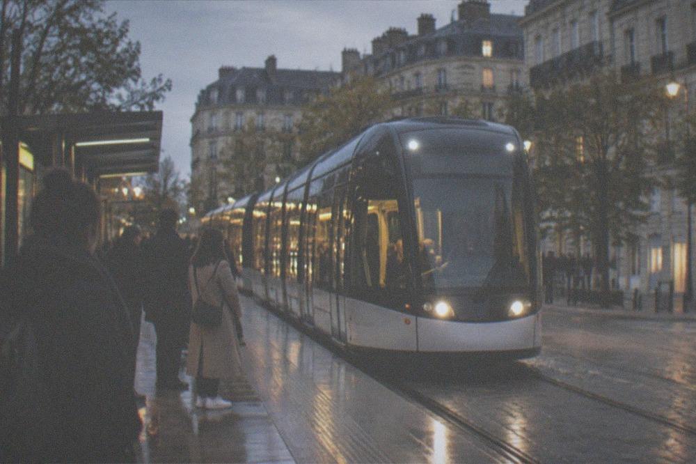 A lit Nantes tram passing through the city centre at night with passengers waiting on the platform