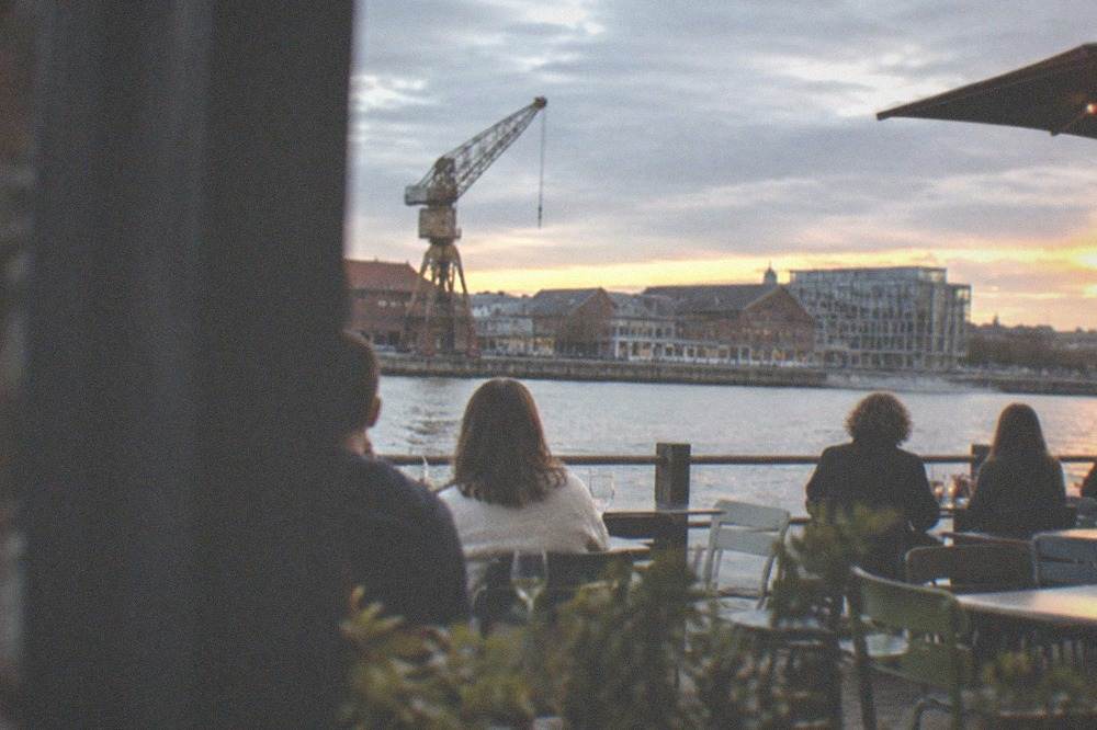 Diners at tables outside a restaurant on the Île de Nantes at dusk, with the Loire beyond