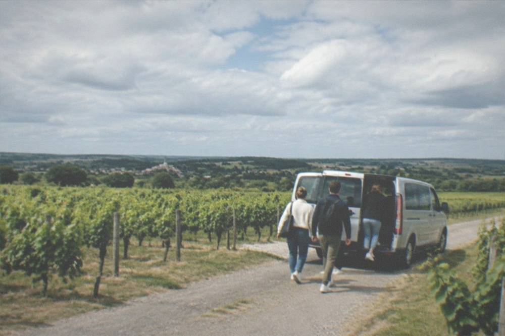 A small van parked on a lane between vineyards with passengers boarding